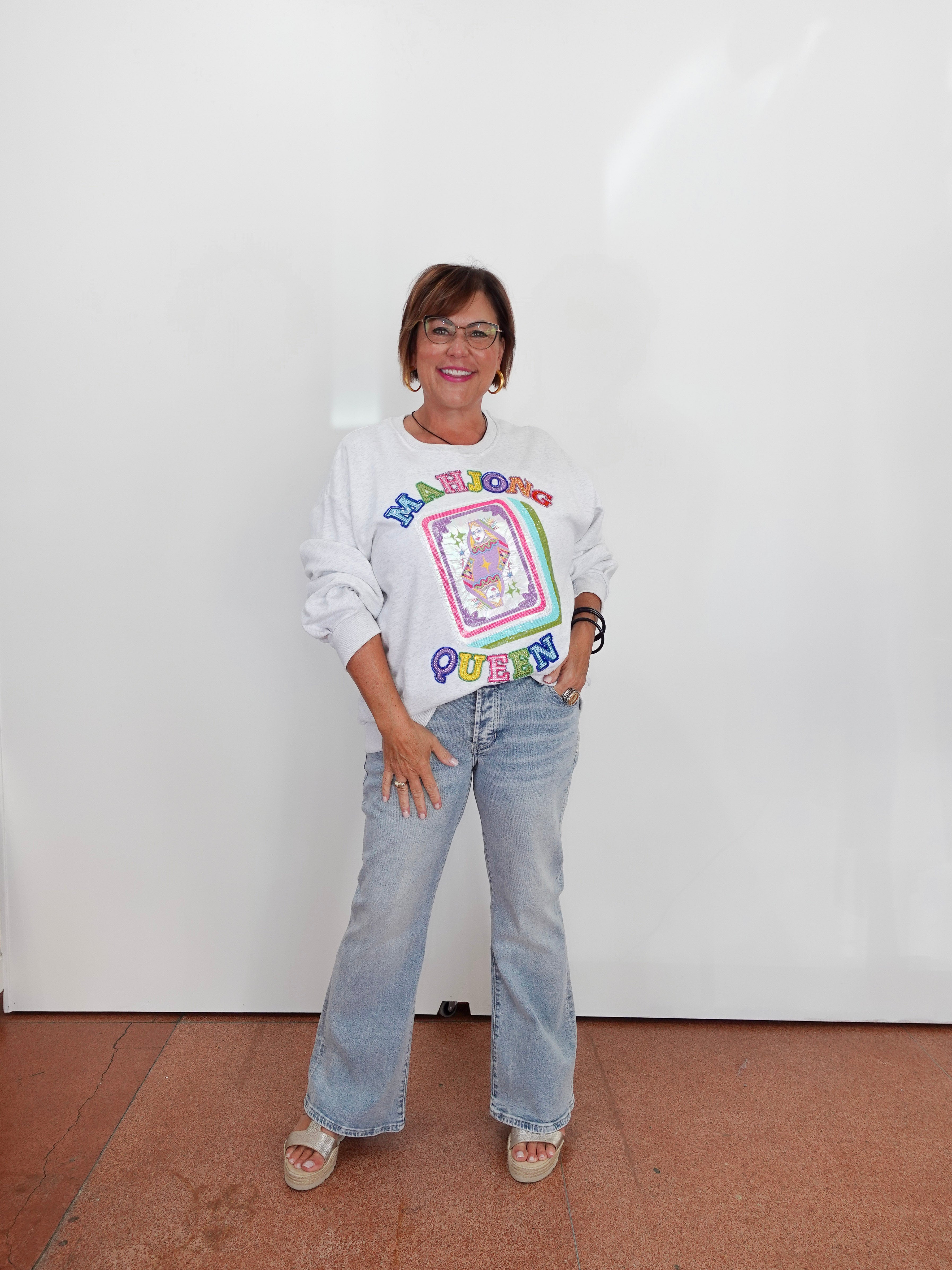A woman stands against a white wall wearing a white long-sleeve graphic sweatshirt with a colorful retro design, light-wash flare jeans, and beige sandals.