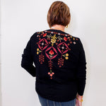 Person wearing a black sweater with colorful embroidery on the back against a white wall.