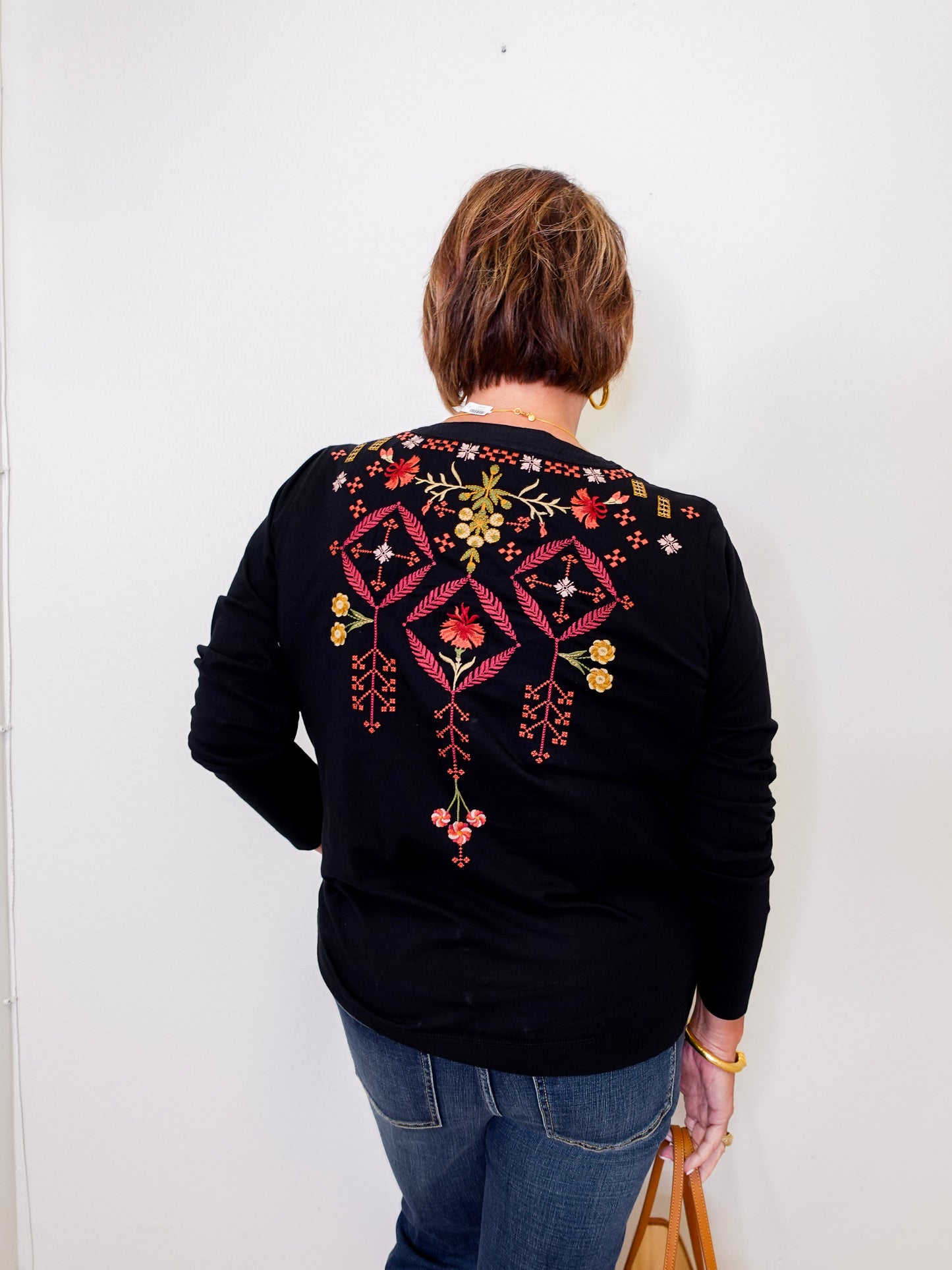 Person wearing a black sweater with colorful embroidery on the back against a white wall.