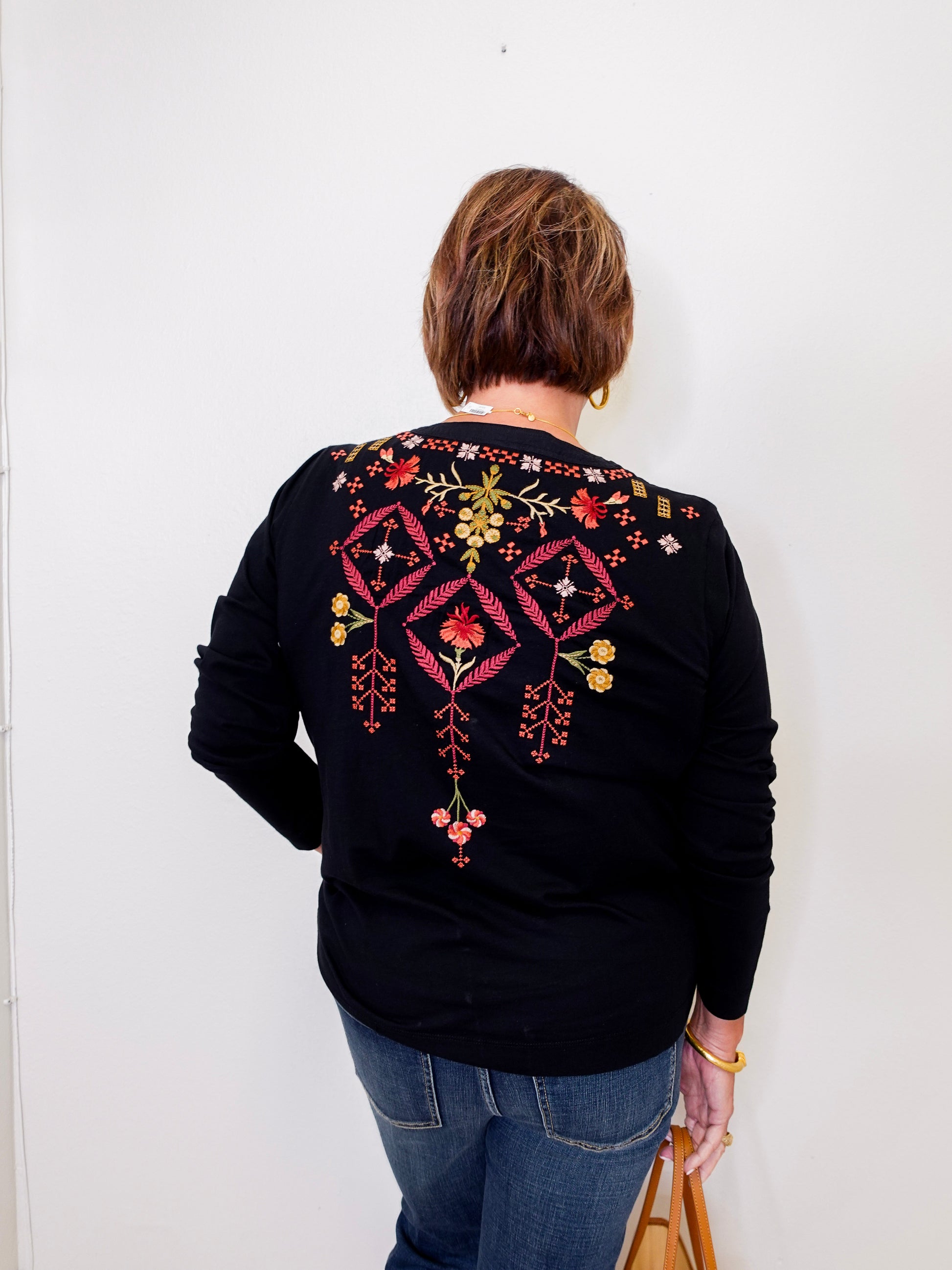 Person wearing a black sweater with colorful embroidery on the back against a white wall.