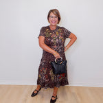 Woman wearing a patterned dress holding a black handbag against a white wall.