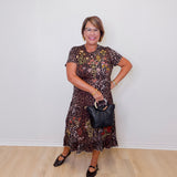 Woman wearing a patterned dress holding a black handbag against a white wall.