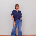 Woman wearing a navy blue top and blue jeans standing against a plain white wall.