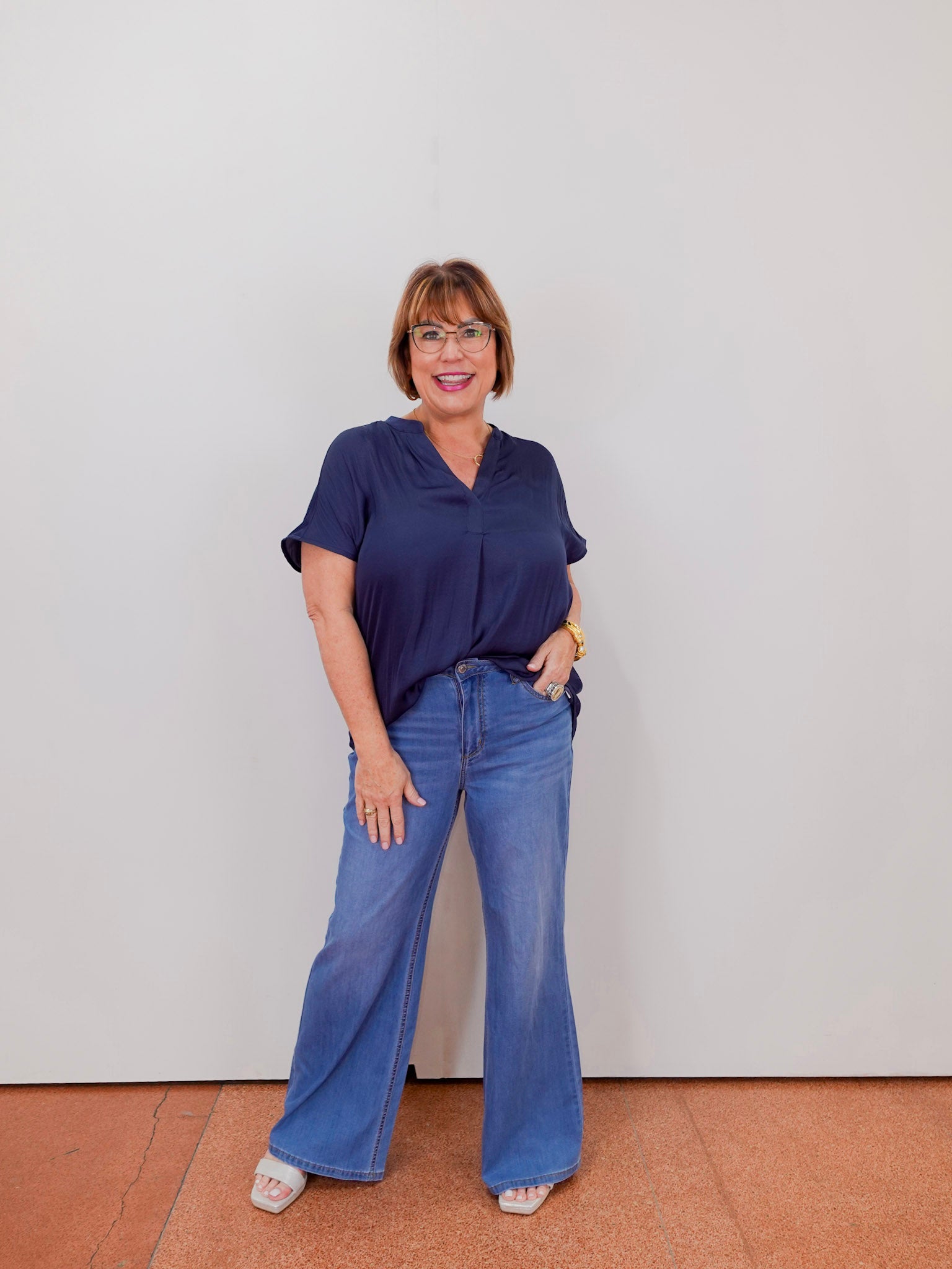 Woman wearing a navy blue top and blue jeans standing against a plain white wall.