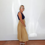 Woman wearing a navy top and beige skirt standing against a white wall.