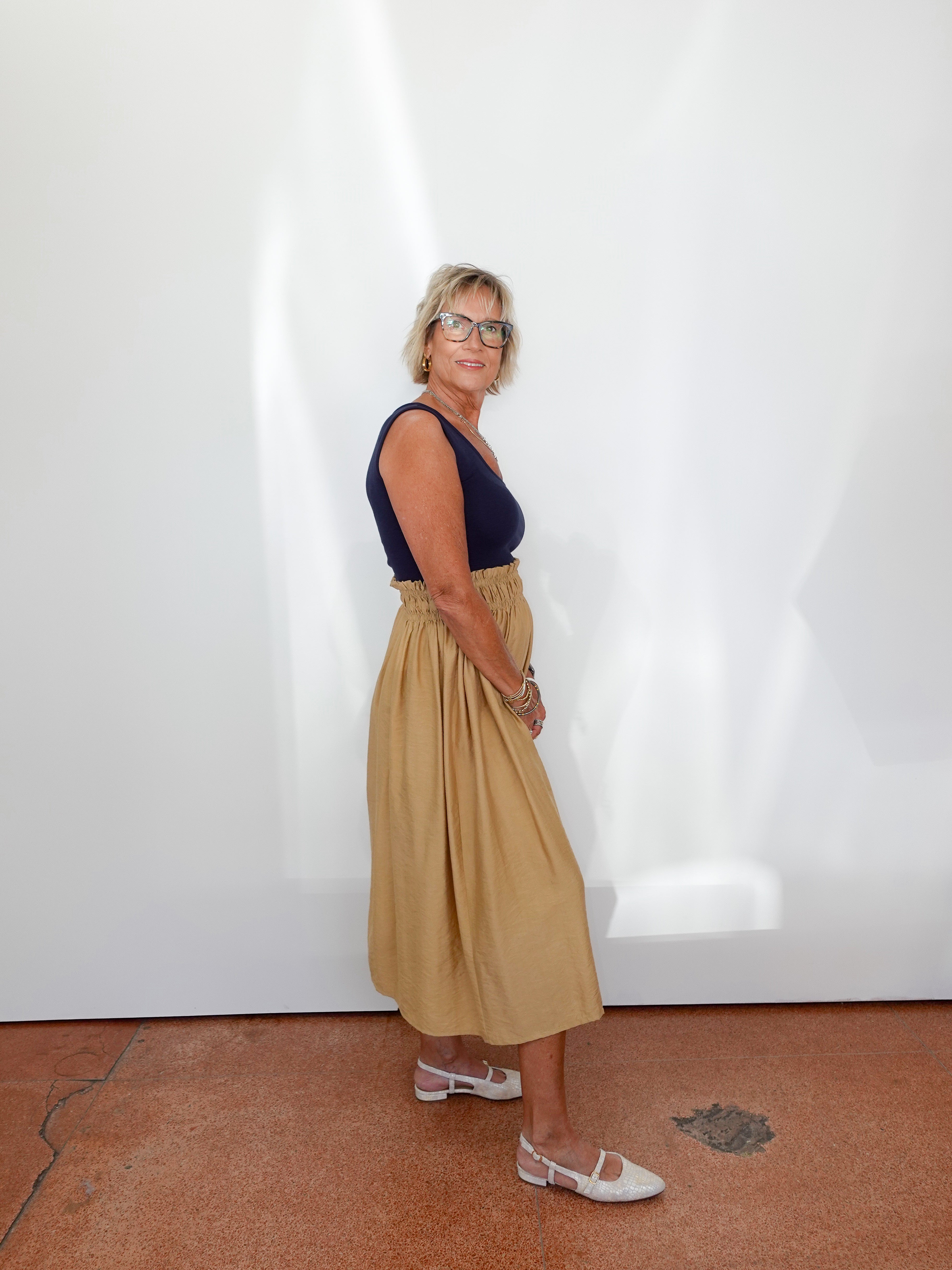 Woman wearing a navy top and beige skirt standing against a white wall.