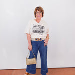 Woman wearing a graphic t-shirt and blue jeans holding a handbag against a white wall.