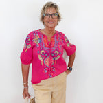 Woman wearing a pink embroidered blouse and beige pants against a white background