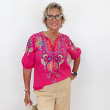 Woman wearing a pink embroidered blouse and beige pants against a white background