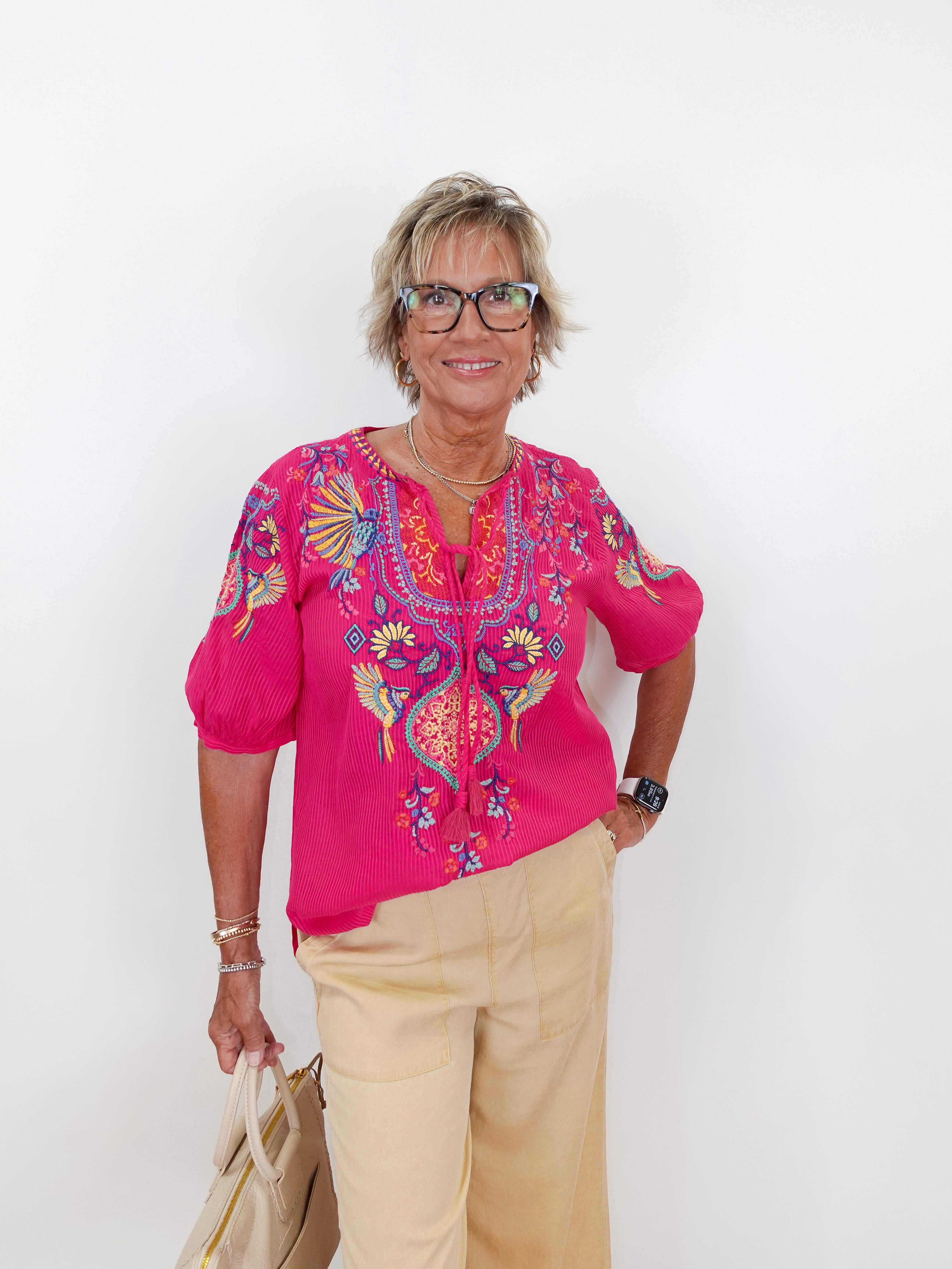 Woman wearing a pink embroidered blouse and beige pants against a white background
