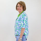 Woman wearing a floral blouse and blue pants against a white wall.