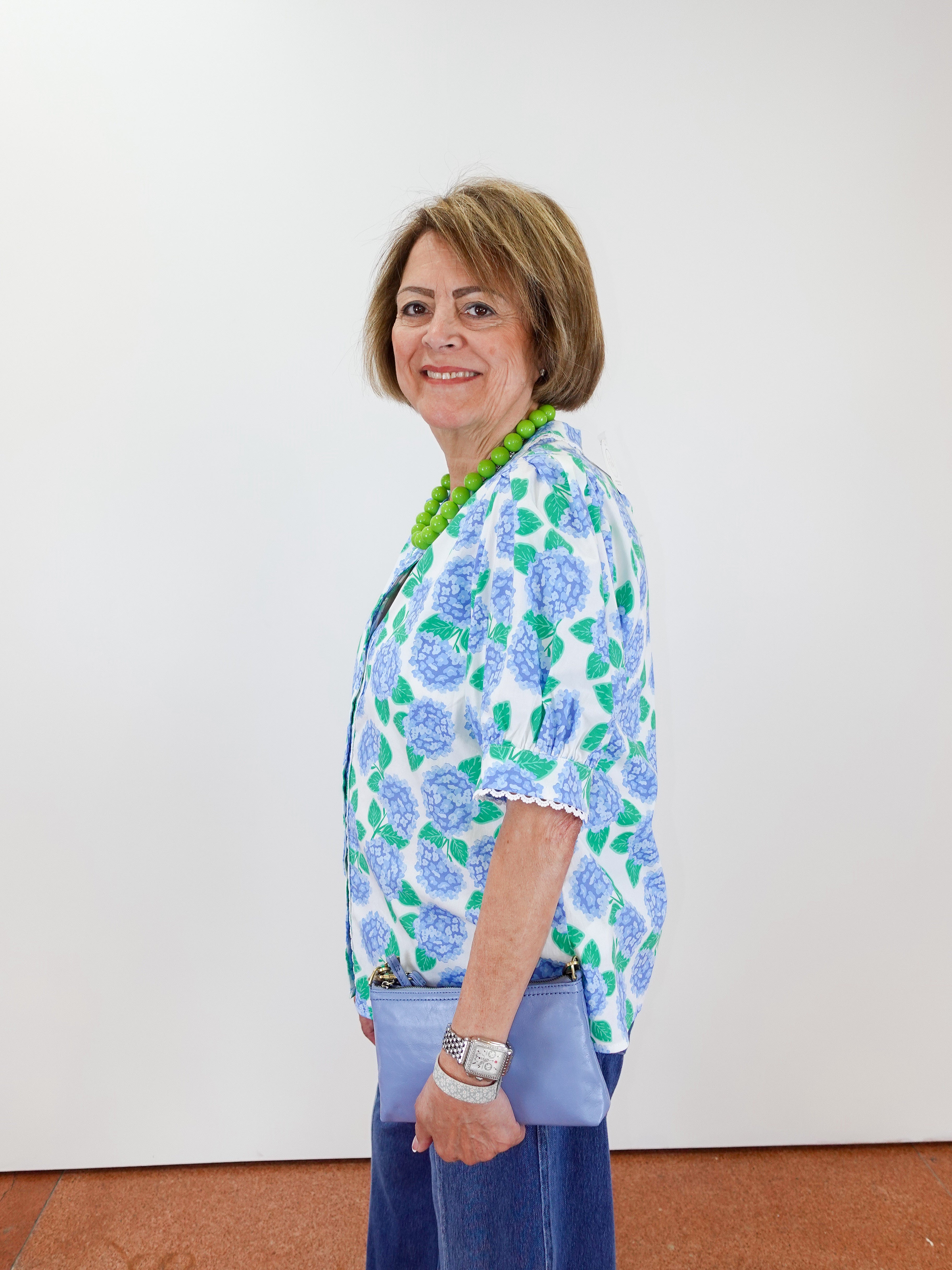 Woman wearing a floral blouse and blue pants against a white wall.