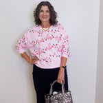 Woman wearing a pink sweater with Christmas lights design, holding a silver handbag against a white wall.