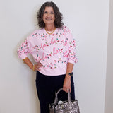Woman wearing a pink sweater with Christmas lights design, holding a silver handbag against a white wall.