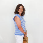 Woman wearing a blue and white striped sleeveless top with white pants, holding a brown bag against a white background.
