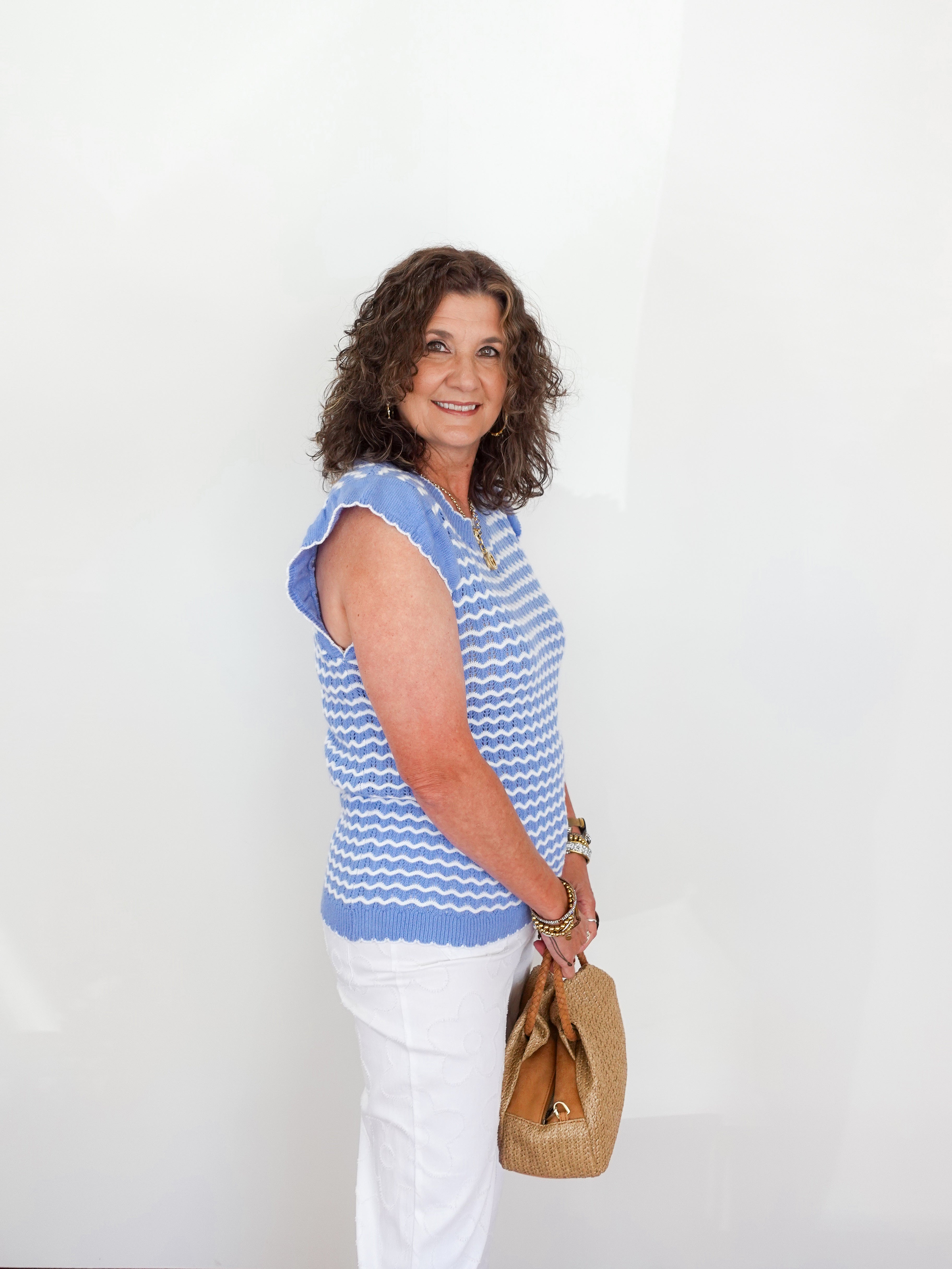 Woman wearing a blue and white striped sleeveless top with white pants, holding a brown bag against a white background.