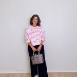 Woman wearing a pink floral blouse and black pants, holding a silver handbag against a white wall.