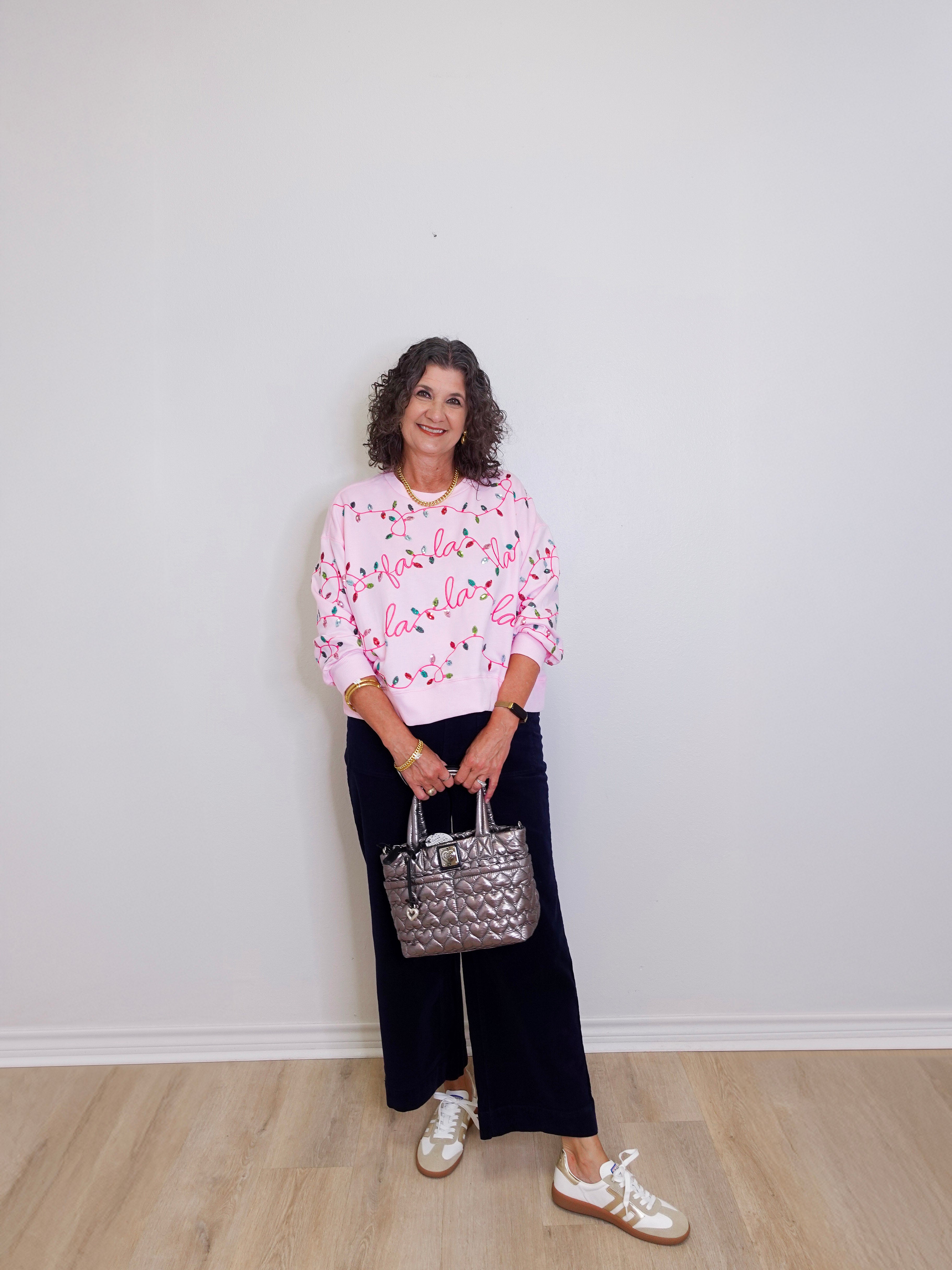 Woman wearing a pink floral blouse and black pants, holding a silver handbag against a white wall.