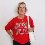 Person wearing a red sweatshirt with 'JOYFUL' text, holding a white bag against a plain background