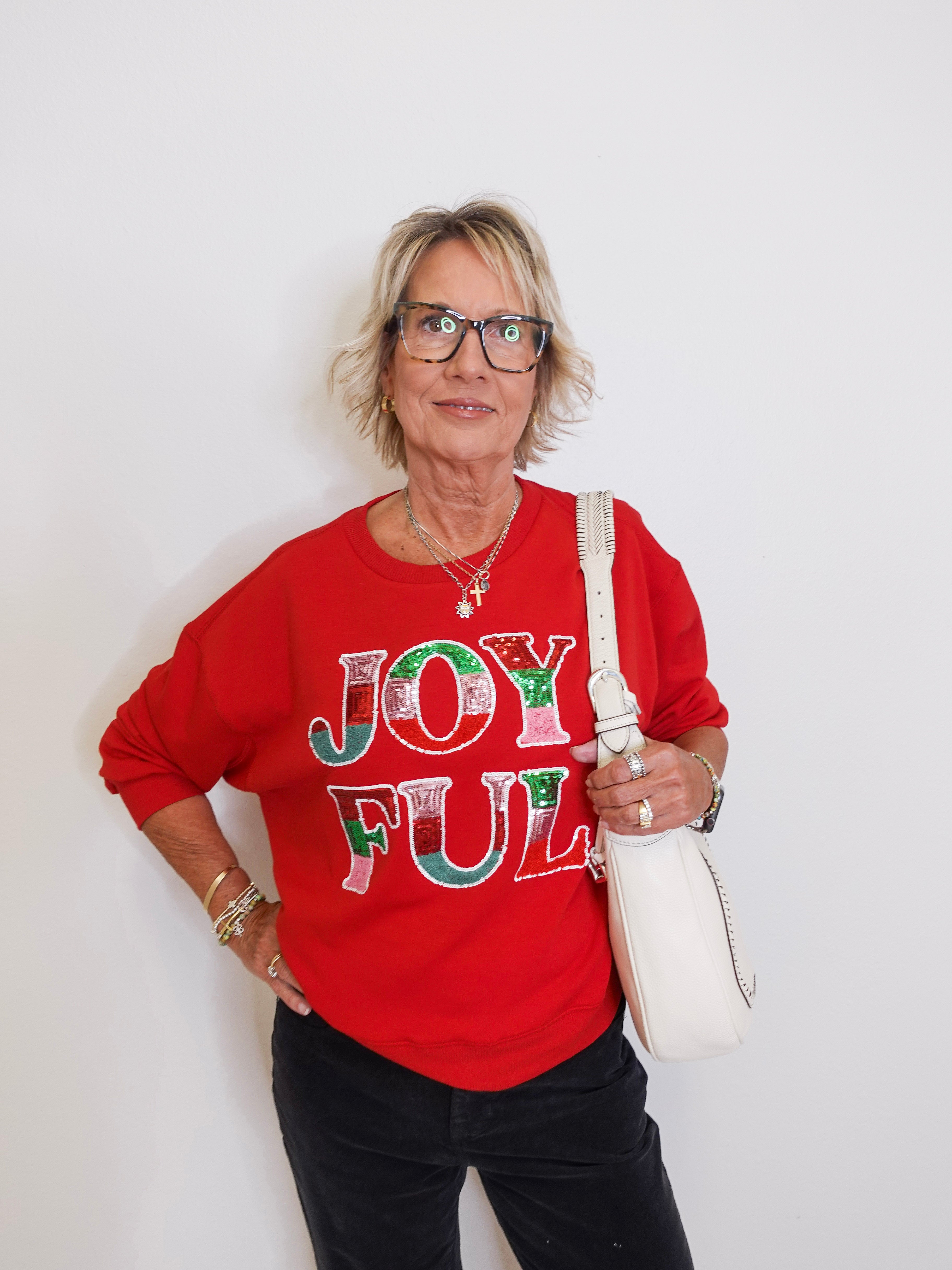Person wearing a red sweatshirt with 'JOYFUL' text, holding a white bag against a plain background