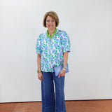 Woman wearing a floral blouse and blue jeans standing against a white wall.