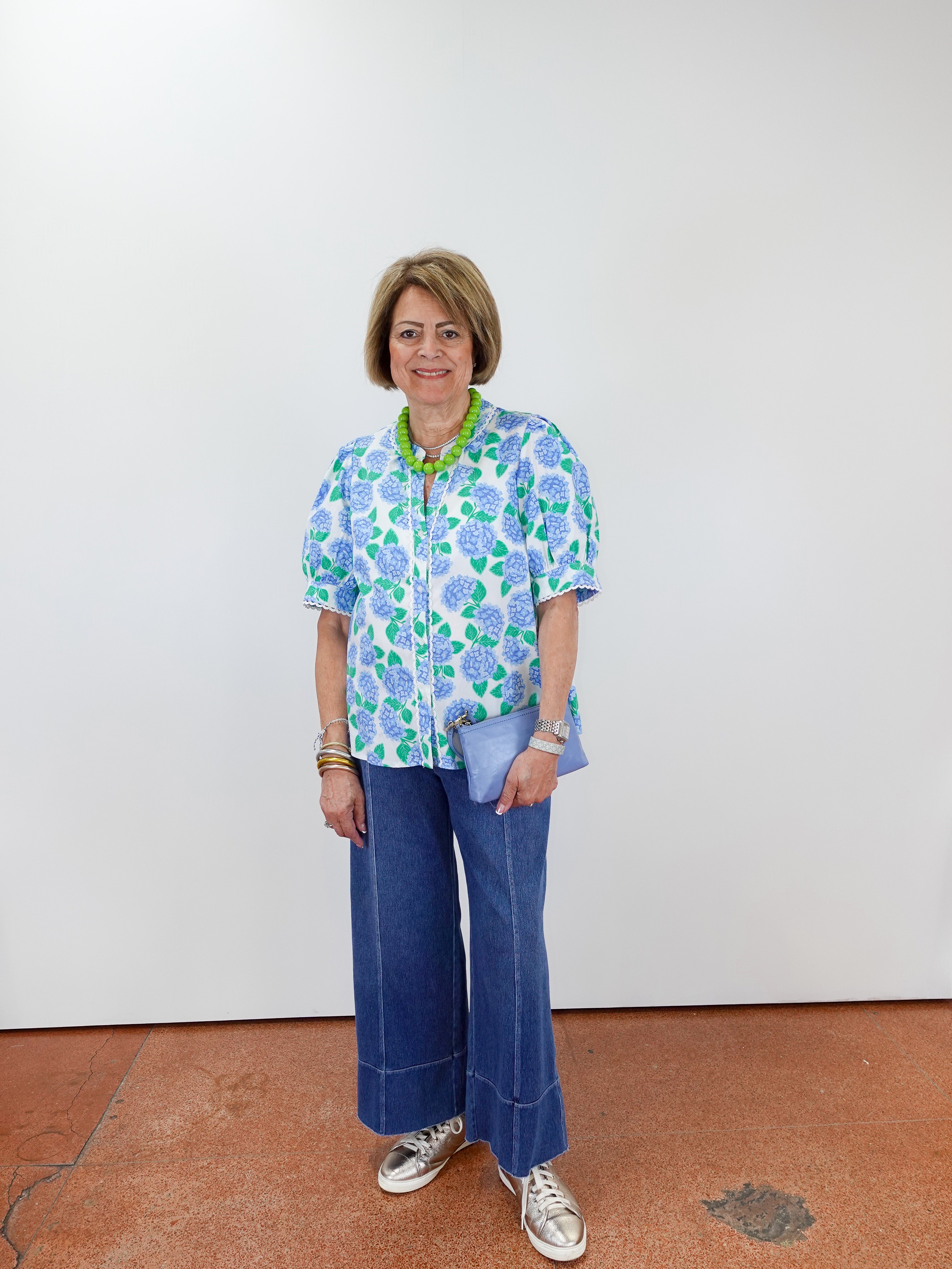 Woman wearing a floral blouse and blue jeans standing against a white wall.
