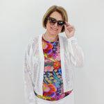 Woman wearing a colorful floral top, white cardigan, and sunglasses against a plain background
