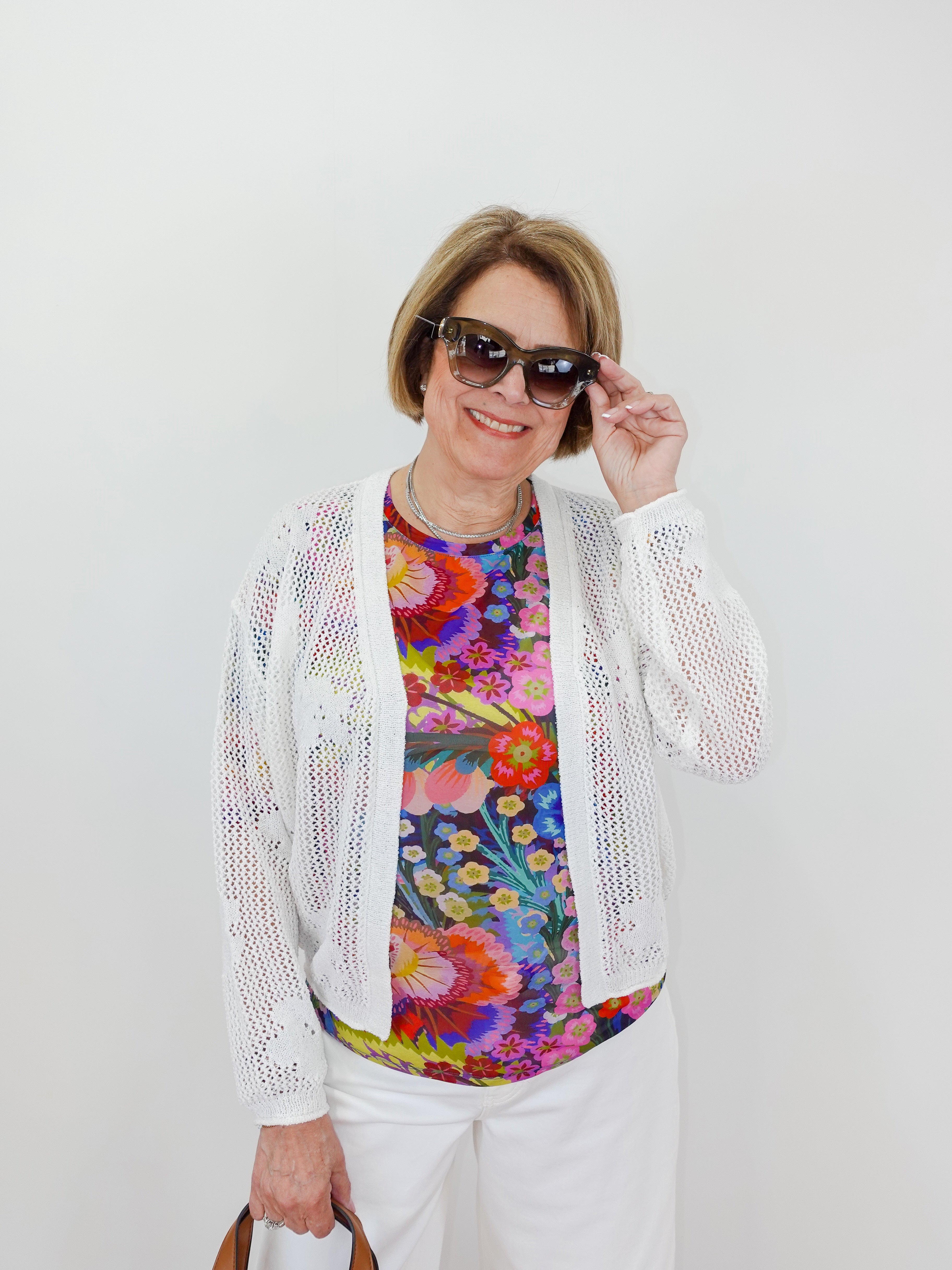 Woman wearing a colorful floral top, white cardigan, and sunglasses against a plain background