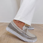 Beige slip-on shoes with rhinestone embellishments worn with white pants on a wooden floor.