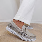 Beige slip-on shoes with rhinestone embellishments worn with white pants on a wooden floor.