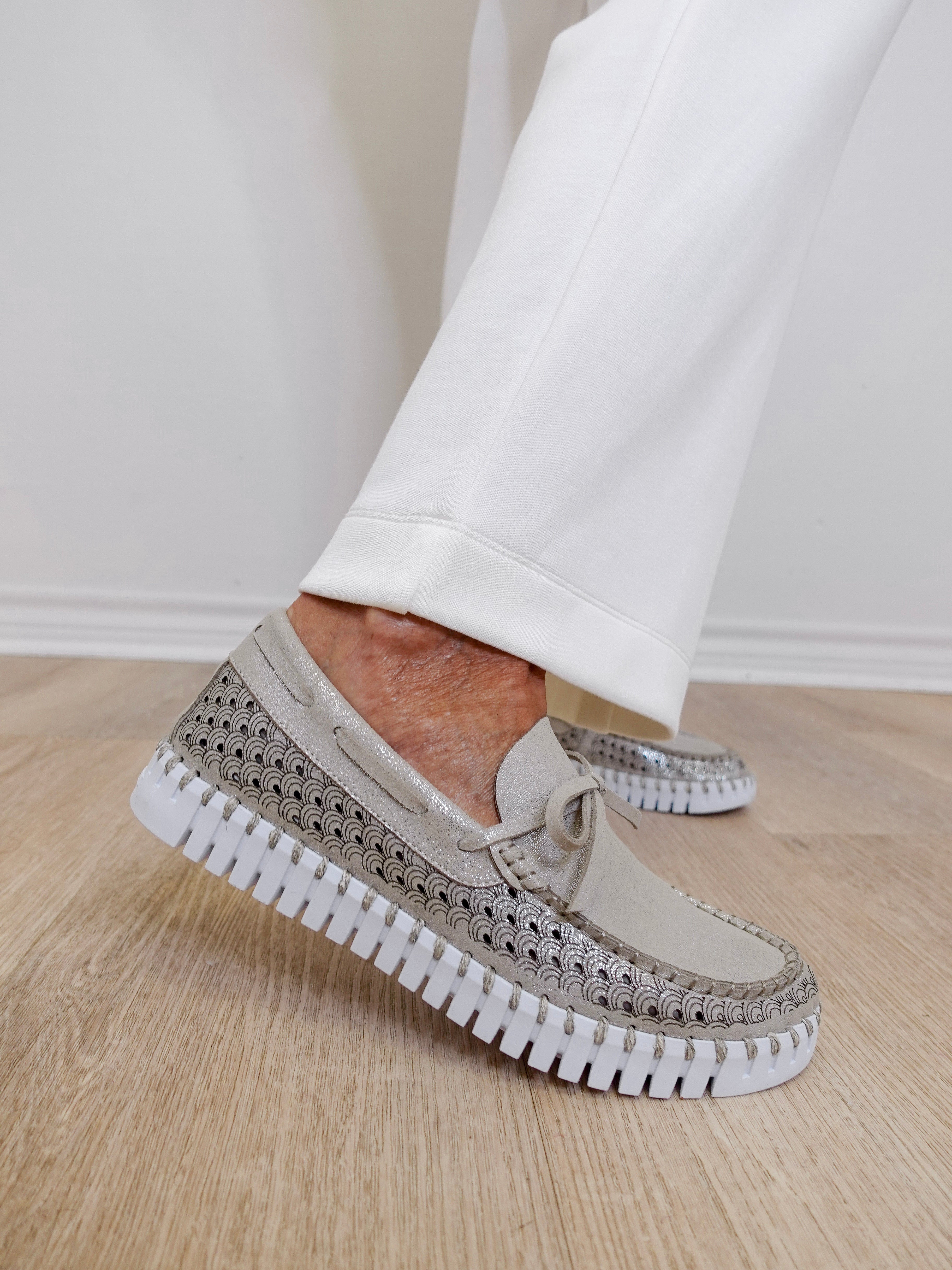 Beige slip-on shoes with rhinestone embellishments worn with white pants on a wooden floor.