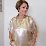 Woman wearing a gold blouse standing against a white wall.