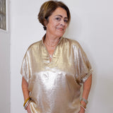 Woman wearing a gold blouse standing against a white wall.