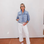 Person wearing a denim jacket, white pants, and pink sneakers against a white wall.