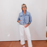 Person wearing a denim jacket, white pants, and pink sneakers against a white wall.