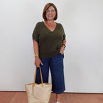 A woman wearing a dark olive green v-neck t-shirt, blue cropped pants, and white slide sandals, holding a beige woven tote bag with tan handles against a plain white wall.