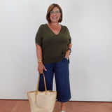A woman wearing a dark olive green v-neck t-shirt, blue cropped pants, and white slide sandals, holding a beige woven tote bag with tan handles against a plain white wall.