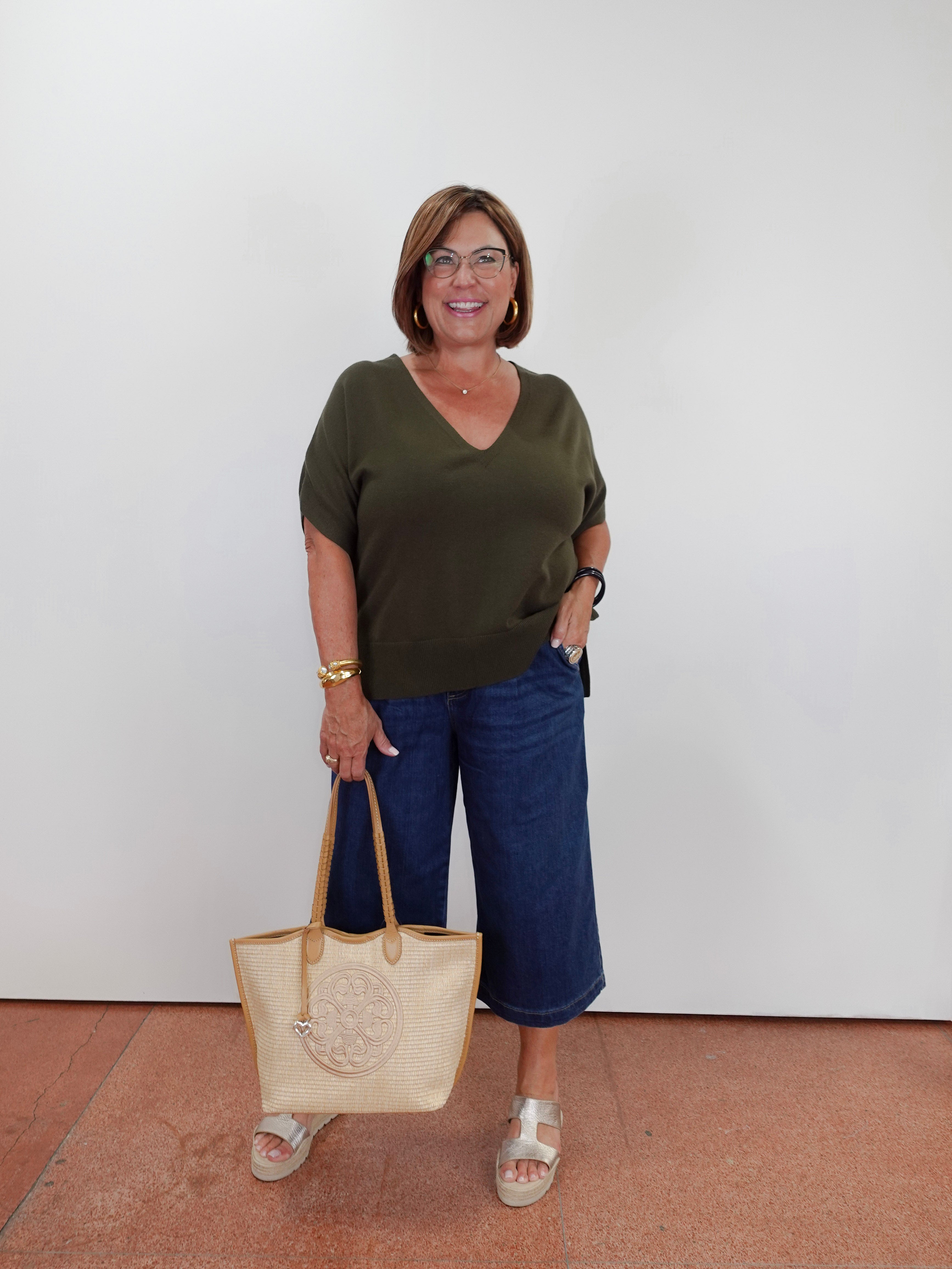 A woman wearing a dark olive green v-neck t-shirt, blue cropped pants, and white slide sandals, holding a beige woven tote bag with tan handles against a plain white wall.