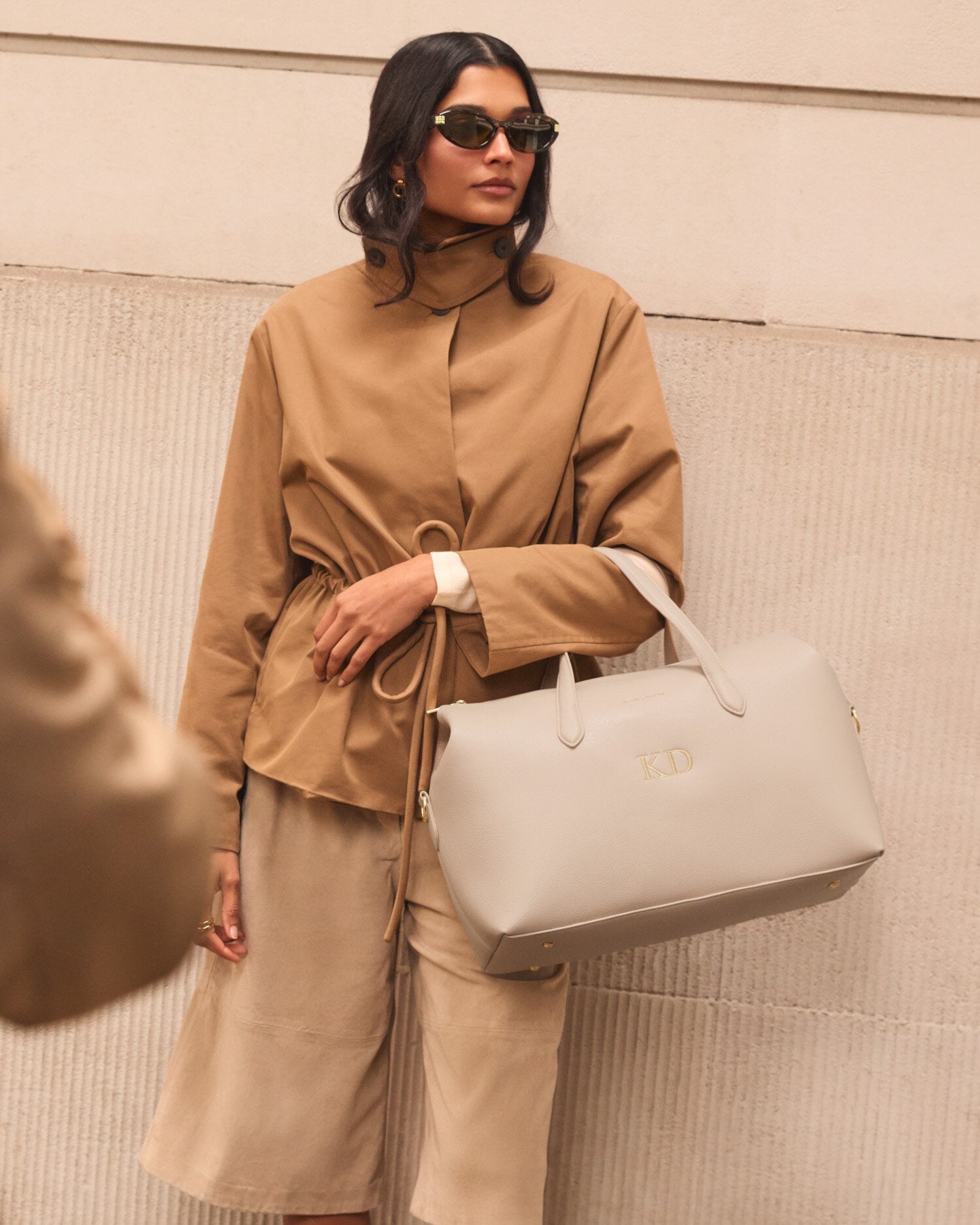 Woman in a beige outfit holding a light gray handbag against a neutral background