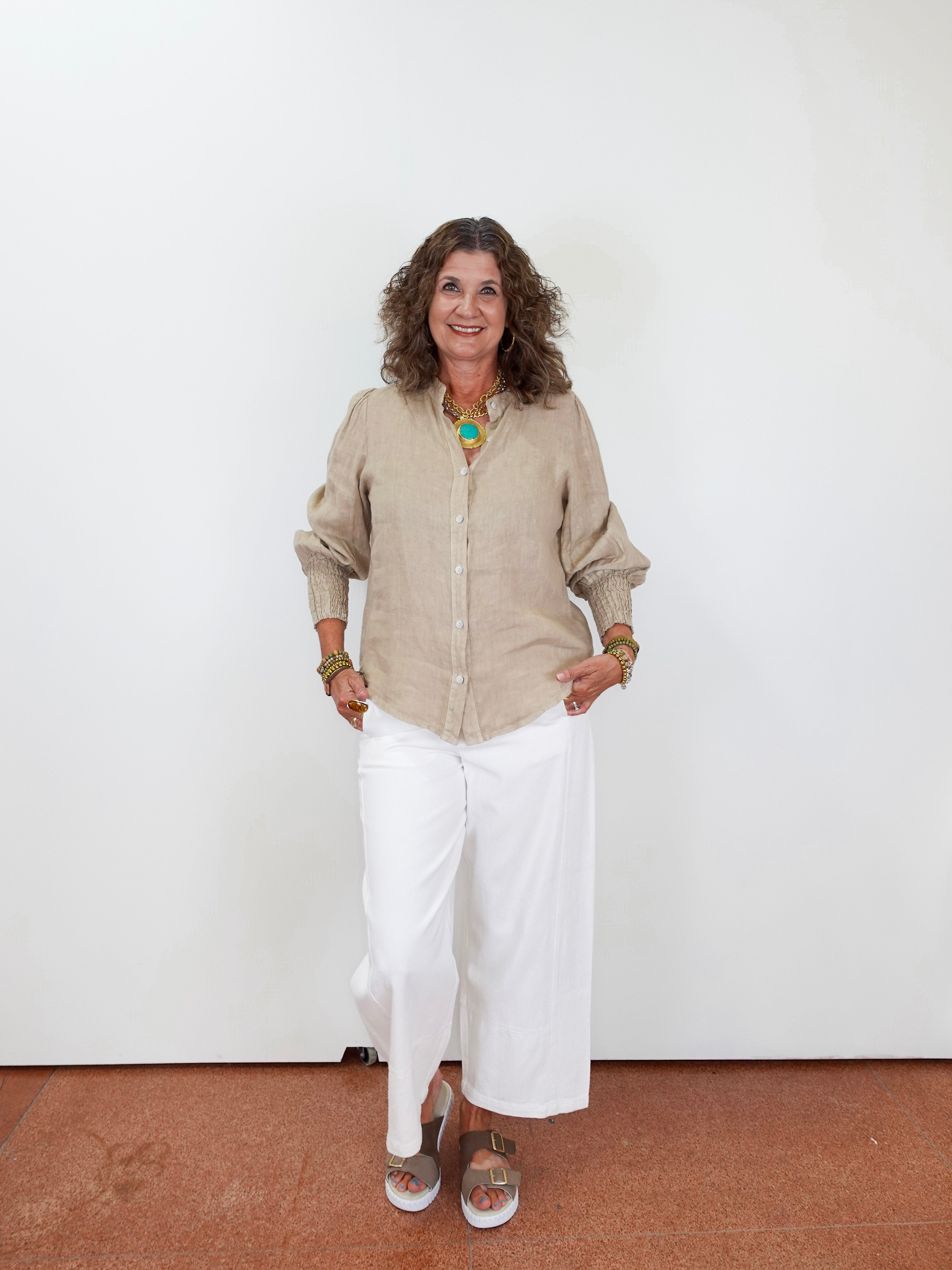 Woman wearing a beige shirt and white pants against a white wall.