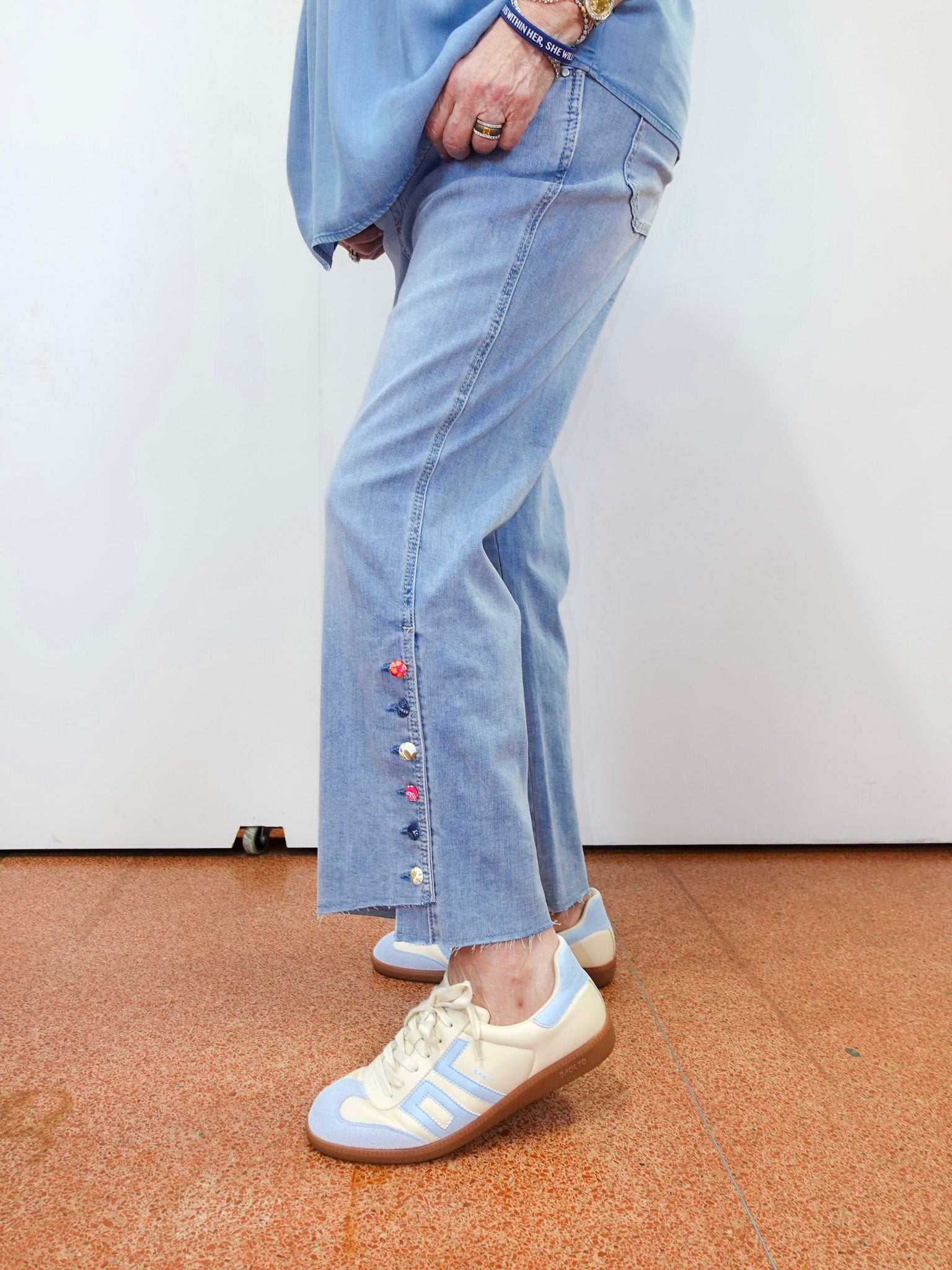 Person wearing light blue jeans with colorful buttons and white sneakers on a plain background