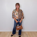Woman wearing a patterned shirt, jeans, and holding a brown handbag against a white wall.