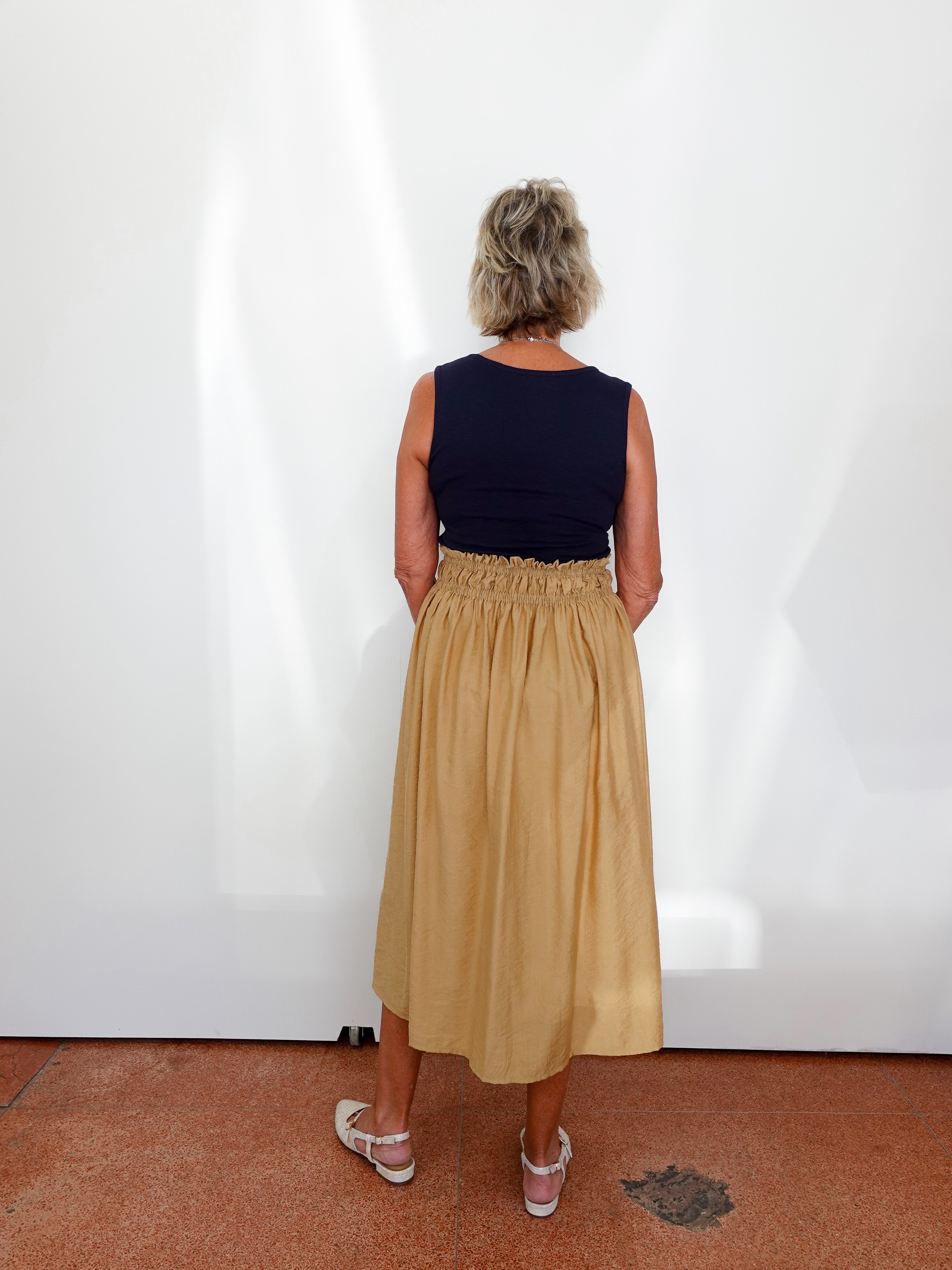 Person wearing a navy top and beige skirt standing against a white wall.