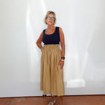 Woman wearing a navy top and beige skirt standing against a white wall.