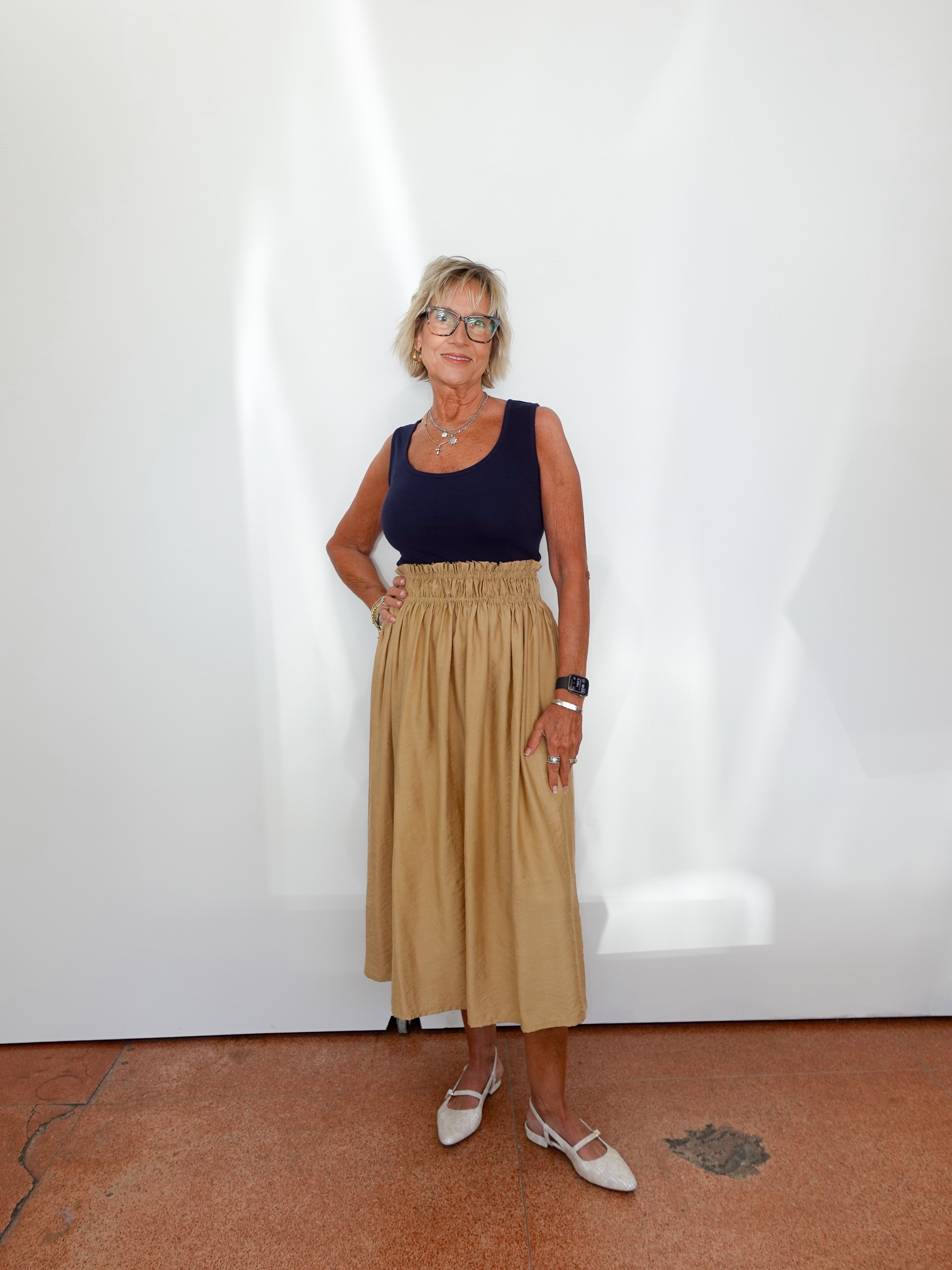 Woman wearing a navy top and beige skirt standing against a white wall.