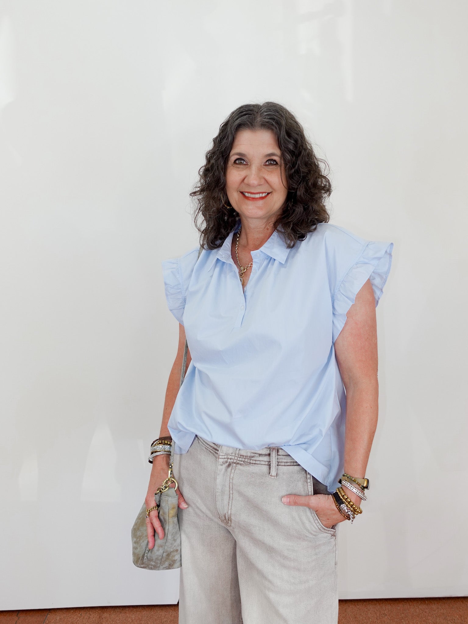 Woman wearing a light blue blouse and beige pants against a white wall