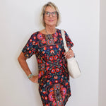 Woman wearing a colorful floral dress holding a white bag against a plain background