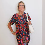 Woman wearing a colorful floral dress holding a white bag against a plain background