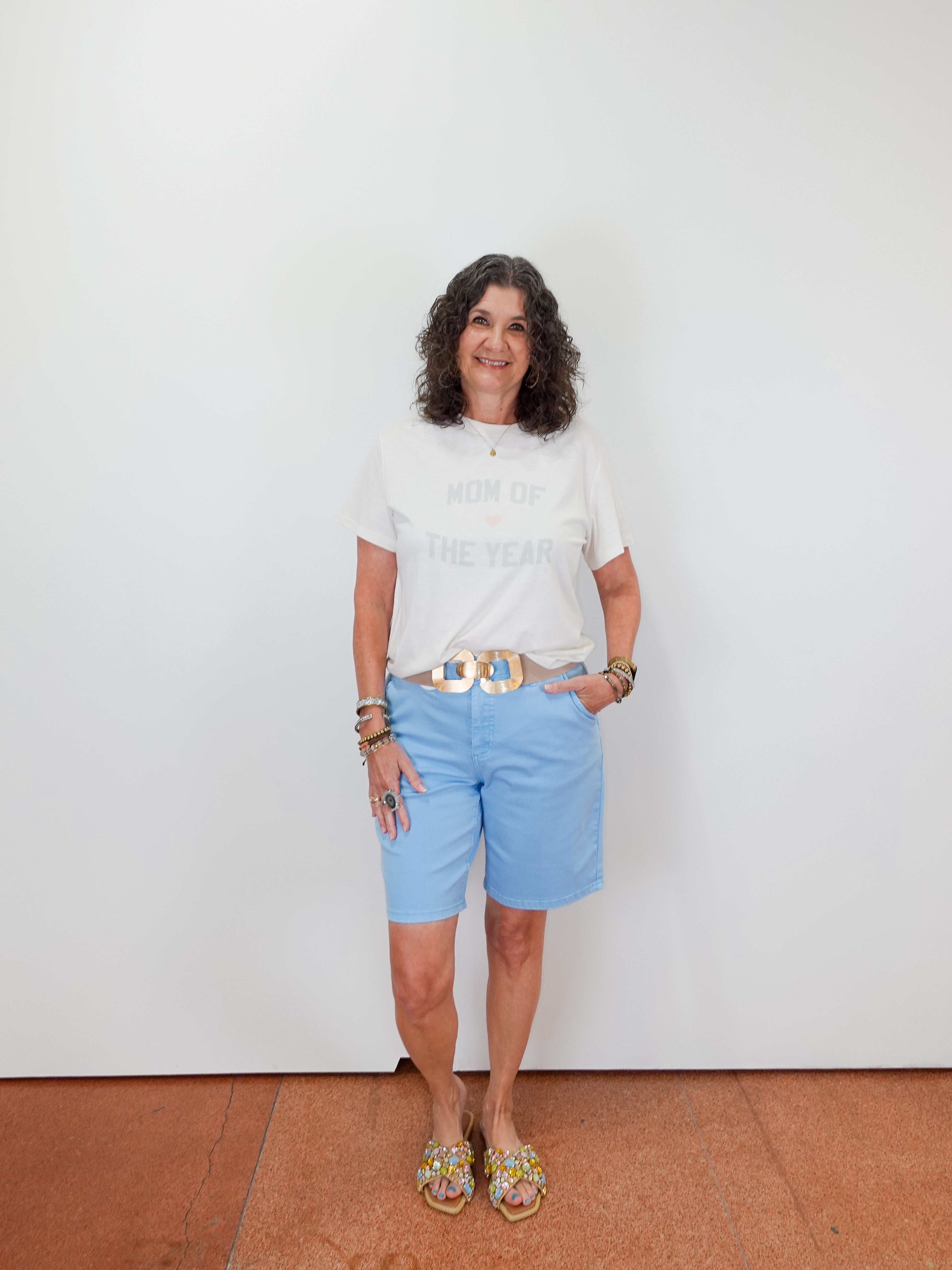 Woman wearing a white t-shirt and light blue shorts standing against a plain white wall.