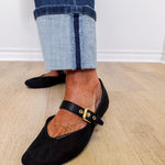 Black flat shoes with a strap worn with rolled-up jeans on a wooden floor.